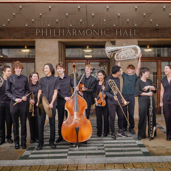 Members of the Youth Company standing in a row outside Liverpool Philharmonic Hall wearing black and holding their instruments