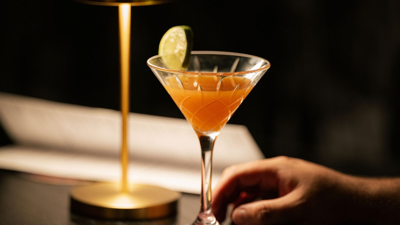 An orange cocktail in a martini glass on the table next to a dim lamp