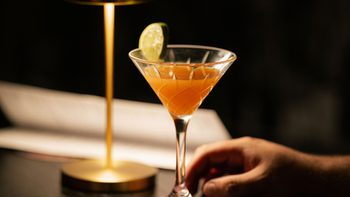 An orange cocktail in a martini glass on the table next to a dim lamp