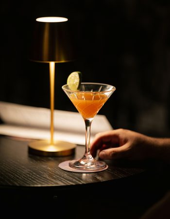 An orange cocktail in a martini glass on the table next to a dim lamp