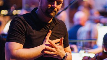 A male BSL interpreter signing, with the Orchestra behind him