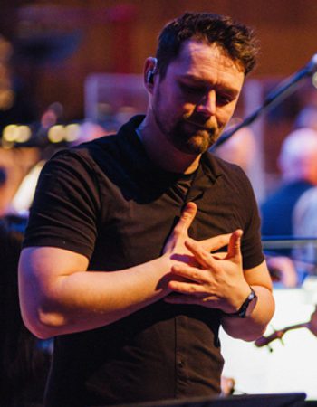 A male BSL interpreter signing, with the Orchestra behind him