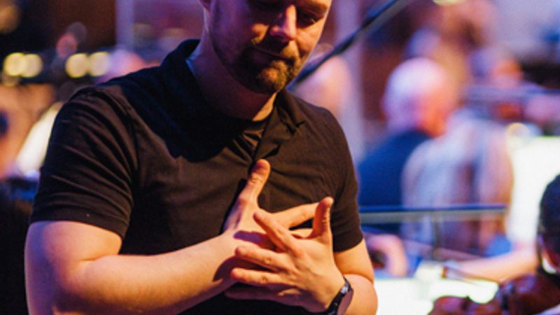 A male BSL interpreter signing, with the Orchestra behind him