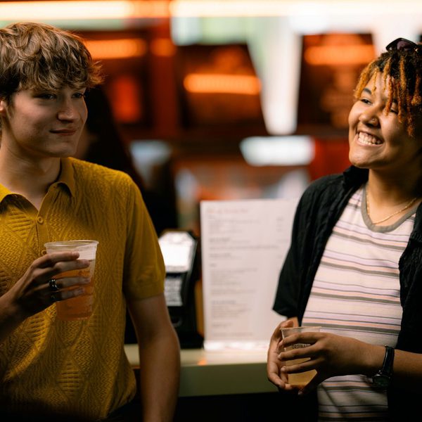 Young man with short brown hair and yellow shirt and a young woman with dreadlocks leaning against a bar holding beers