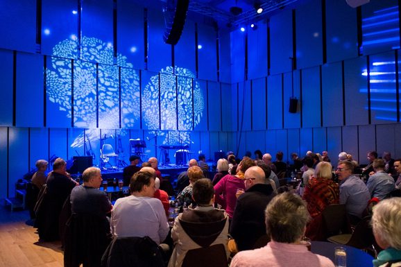 Photo of Music Room stage with blue background, and audience members seated waiting for a performance to start