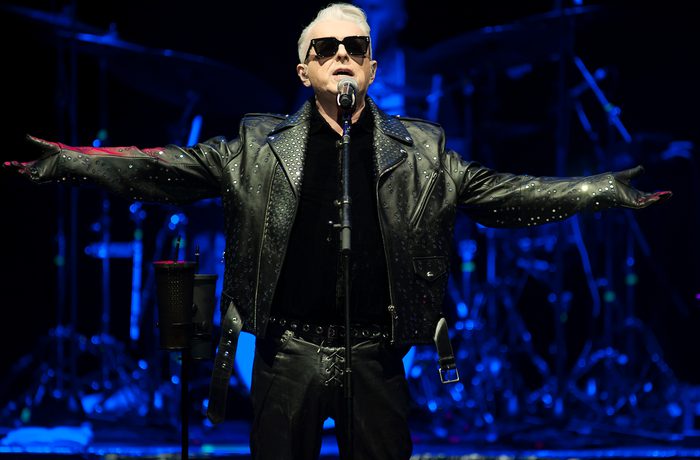 Holly Johnson, a white man with short grey hair and black sunglasses, wearing a leather jacket and singing on stage with his arms outstretched