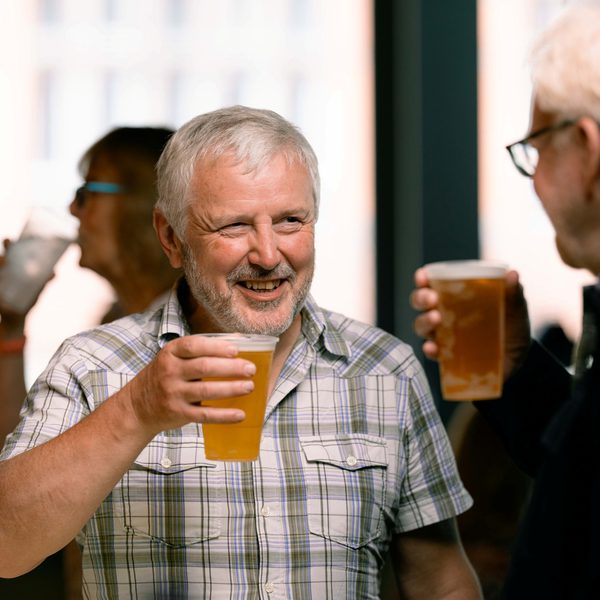 A white man with short grey hair and a grey checked top holding a pint of beer talking to his friend