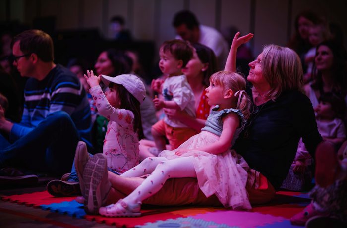 A young girl dressed in a tutu sat on her mother's lap with her arm in the air