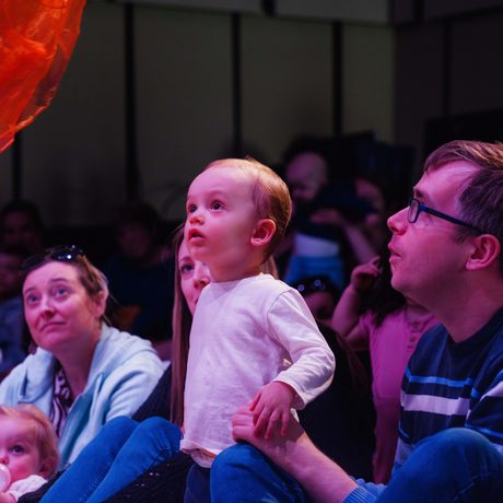A young child looking at a floating piece of red fabric being held up by their father