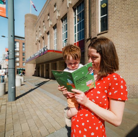 A mother with short brown hair and a red patterned dress, holding her young child reading a family brochure outside Liverpool Philharmonic hall