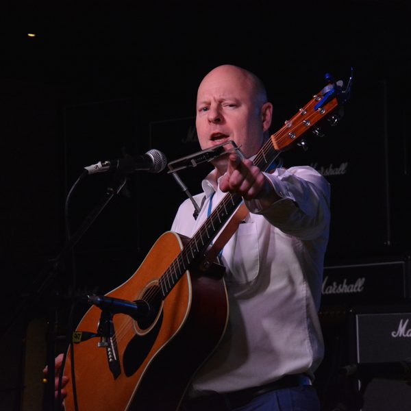 White bald man in white shirt with acoustic guitar, pointing at audience