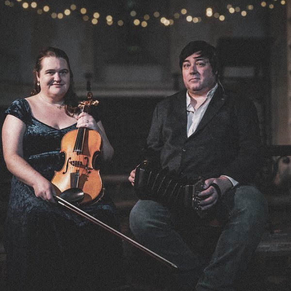 White woman in dark green evening dress with violin, next to a white man in a blazer and jeans with an accordion.