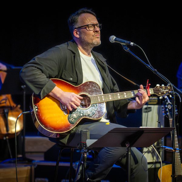 White man in green shirt over white t-shirt, with glasses, playing acoustic guitar.