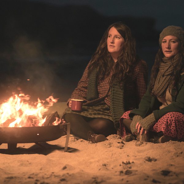 Two women in scarves, one in a beanie, in front of a campfire