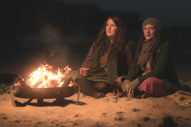 Two women in scarves, one in a beanie, in front of a campfire
