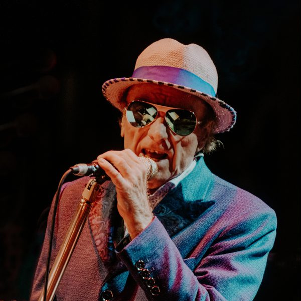 Van Morrison singing into a microphone in front of a black background wearing a cream hat