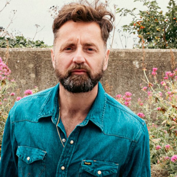 Blue Rose Code, who has short brown hair, wearing a blue denim jacket in a field of flowers