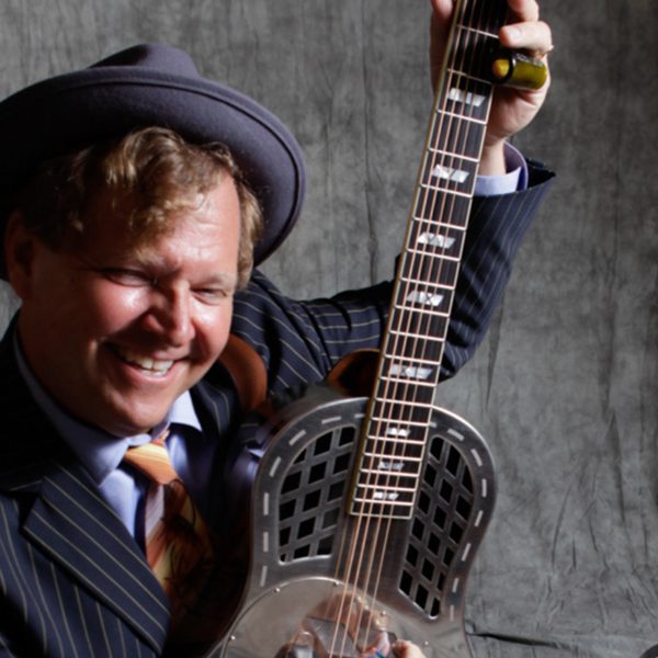 Catfish Keith wearing a grey suit and hat smiling and playing a silver guitar