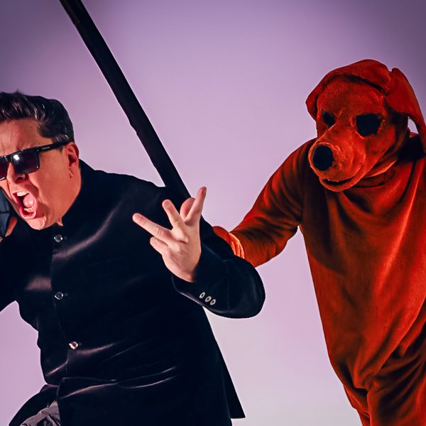 Dom Joly shouting on the phone wearing a black jacket and sunglasses with a person dressed in a red dog costume standing behind him attempting to hit him with a black stick