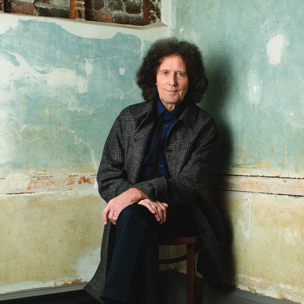 Gilbert O'Sullivan sitting on a wooden stool wearing a black coat looking at the camera in front of a cream and light blue wall