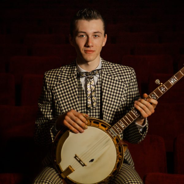 Jake Vaadeland looking into the camera smiling wearing a brown gingham suit holding a banjo guitar in front of a black background