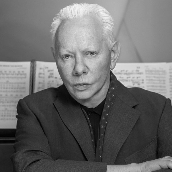 Black and white photo of Joe Jackson looking at the camera in front of white music sheets