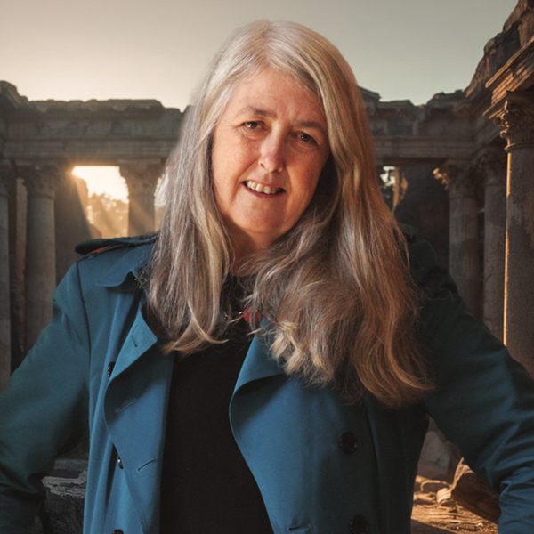 Mary Beard, with long blonde hair, wearing a blue jacket standing in an ancient historic building