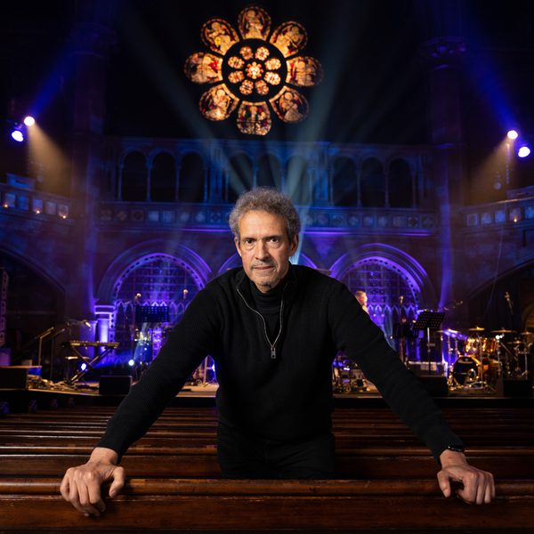 Mike Lindup wearing black and looking into the camera, standing in a church hall with blue hue lighting in the background