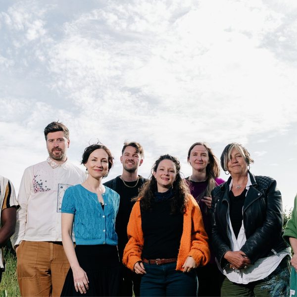 8 members of Spell Songs stood in a line in a field with a cloudy sky above them