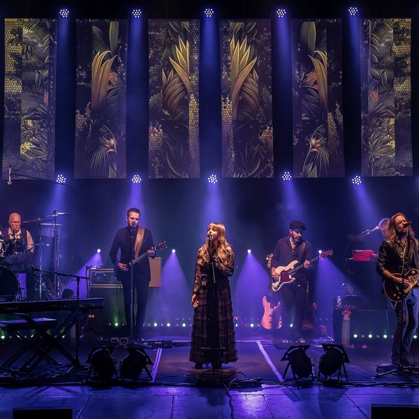 6 members of Rumours of Fleetwood Mac performing in a line on stage with purple lighting
