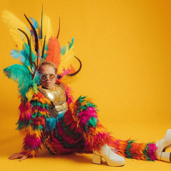 Richard Reeves, a white man with short blonde hair, dressed as Elton John in a feathered multicoloured jumpsuit and headpiece, sat in front of a bright yellow background