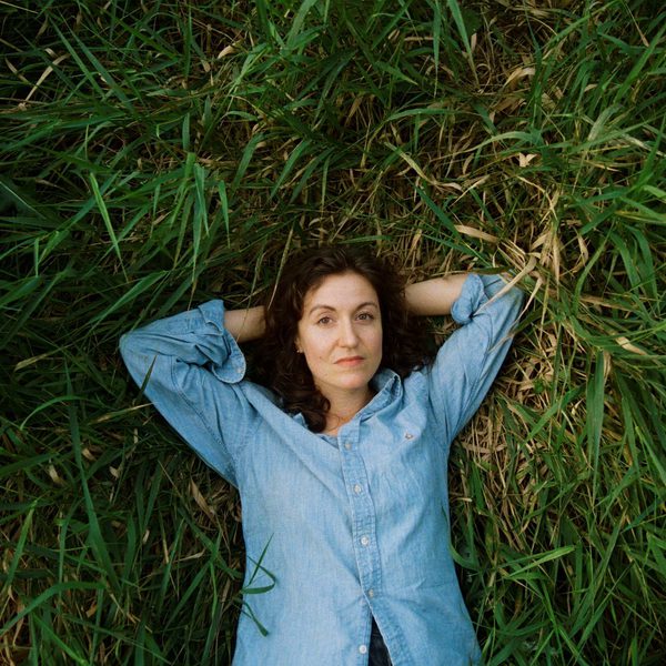 Tessa Rose Jackson, a white woman with curly brown hair, lying in a field with her hands behind her head wearing a blue denim shirt