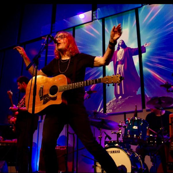 Vinny Peculiar, a white man with shoulder length ginger hair and glasses, performing on the Music Room stage with his arms outstretched