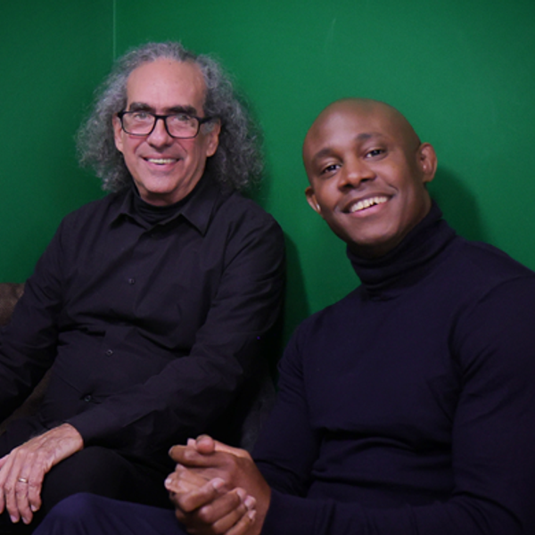 Eduardo Martin, a white man with long grey hair, and Ahmed Dickinson, a bald black man, wearing all black and sat in front of a dark green wall