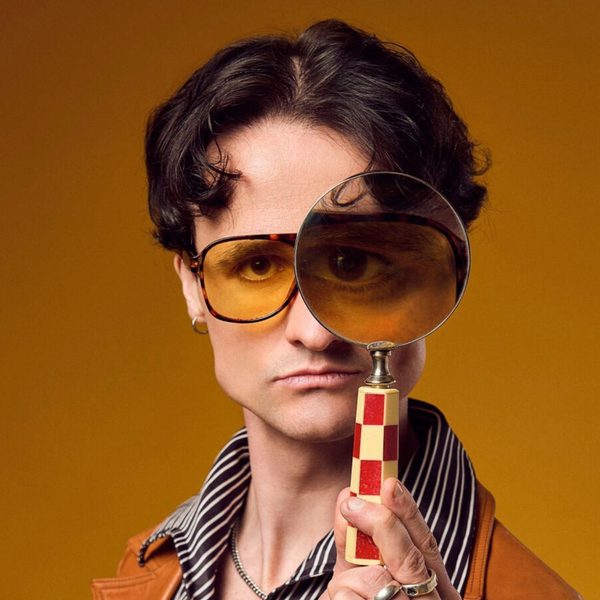 Jazz Emu, a white man with short brown hair and yellow round glasses, looking at the camera through a magnifying glass