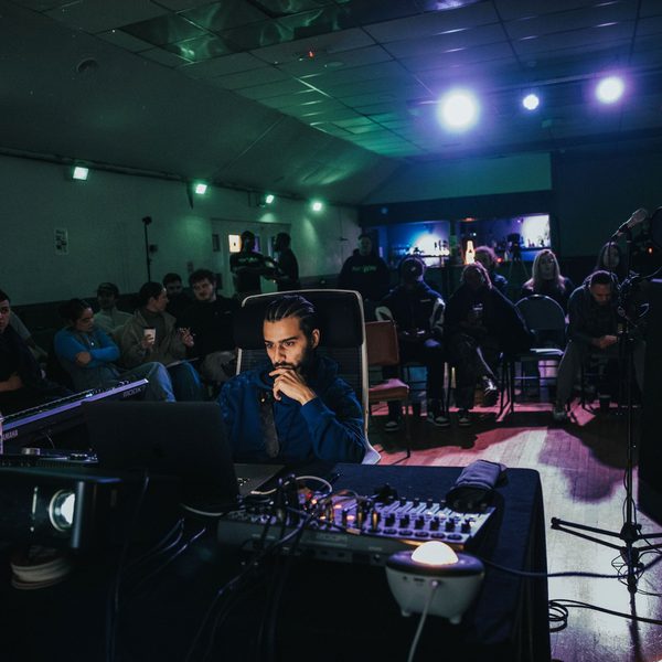 A man sat at a mixing desk being watched from behind by an audience