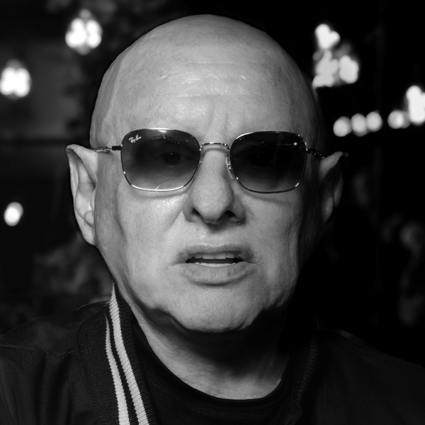 Black and white image of Shaun Ryder, a bald white man, wearing sunglasses and looking directly at the camera