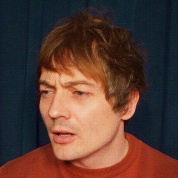 Matt Bragg, a white man with brown hair looking towards the left with a confused expression, wearing a dark orange jumper