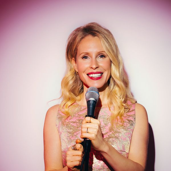 Lucy Beaumont, a white woman with long blonde hair smiling at the camera and holding a microphone