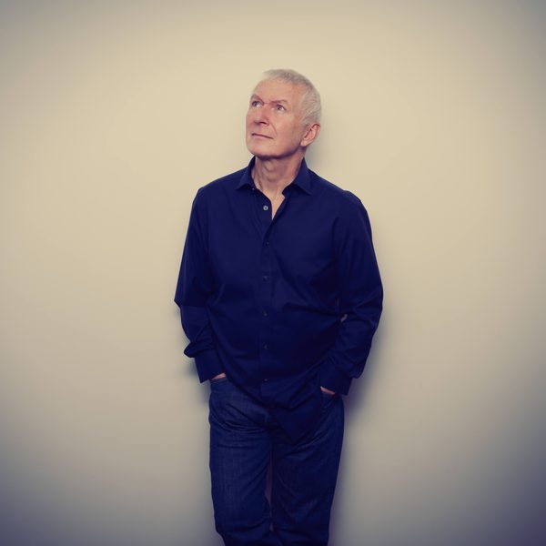 Danny Bowes, a white man with grey hair wearing a dark shirt and looking at the upper left corner and standing in front of a white background