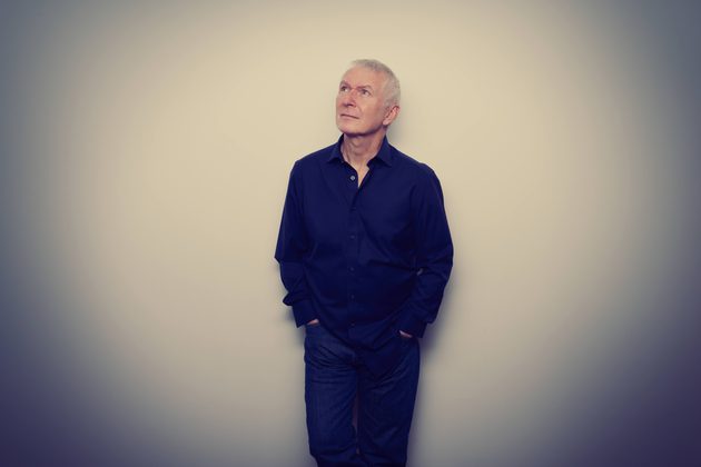 Danny Bowes, a white man with grey hair wearing a dark shirt and looking at the upper left corner and standing in front of a white background