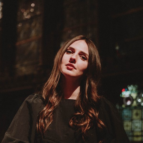 Amelia Coburn with long brown hair, standing against a dark background, and looking at the camera