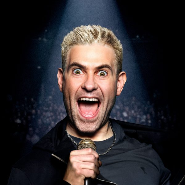 Simon Brodkin with bleached hair, standing a dark background, shouting into a microphone