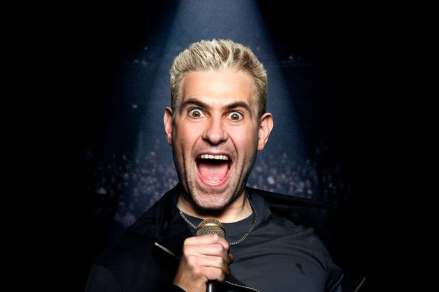 Simon Brodkin with bleached hair, standing a dark background, shouting into a microphone