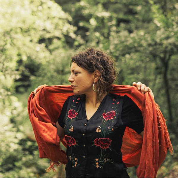 Amy Scott Samuel standing in a woodland, wearing a black dress with red flowers, and holding aloft a red shawl