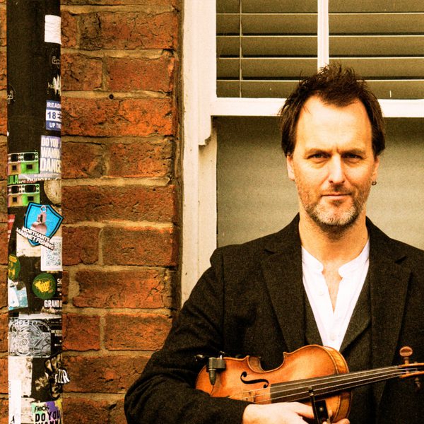 Jon Boden holding a violin, wearing a dark suit and white shirt, standing next to a lamppost in front of window.