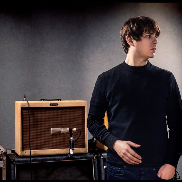 Jake Bugg wearing a black jumper looking to the right and standing in front of a brown guitar amp