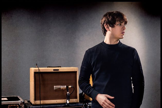 Jake Bugg wearing a black jumper looking to the right and standing in front of a brown guitar amp