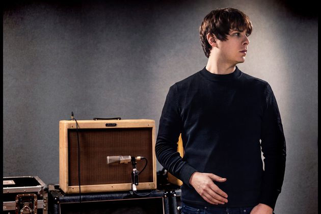 Jake Bugg wearing a black jumper looking to the right and standing in front of a brown guitar amp