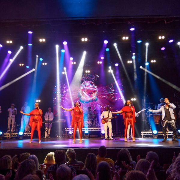 Five singers on stage singing with coordinated hand movements. 3 black women in red outfits and 2 black men in sparkly suits.